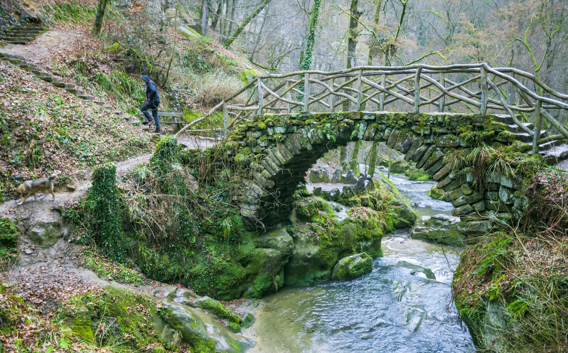 Небольшой арочный каменный мост пересекает ручей, и турист идет по тропе через лес.